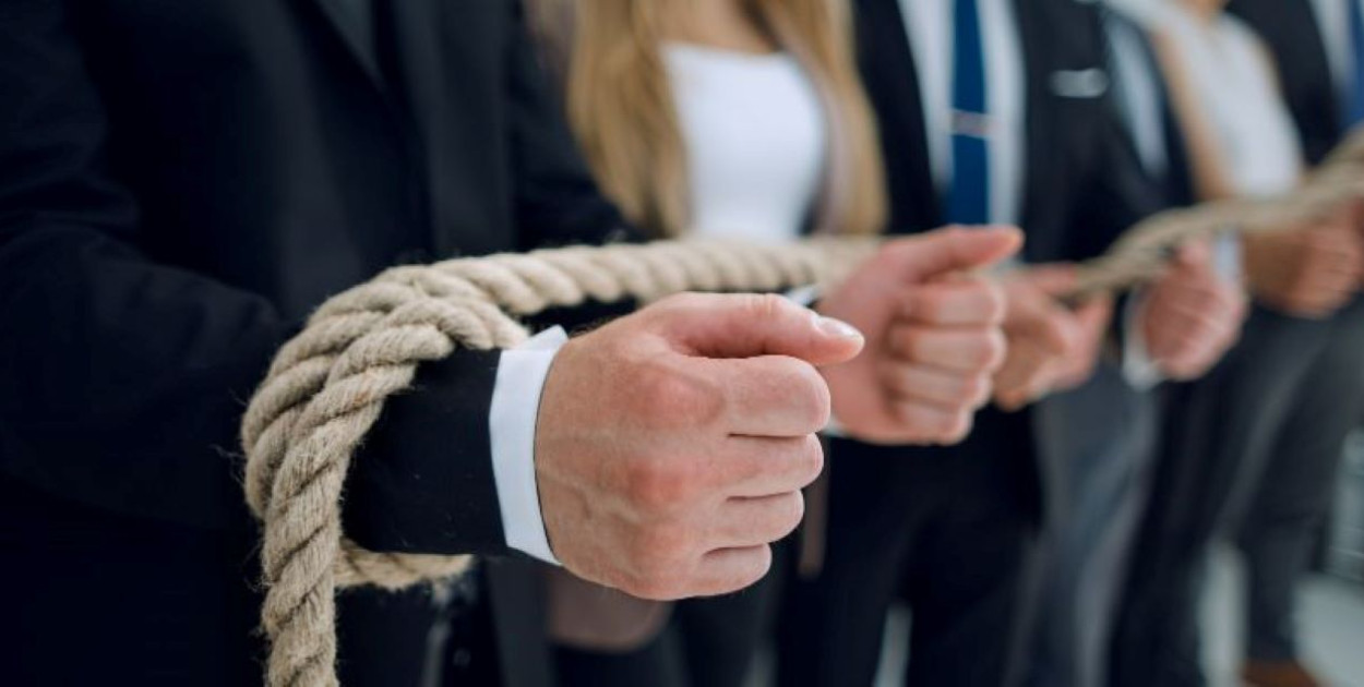 Osoby w formalnych ubraniach stoją w szeregu z grubym sznurem owiniętym wokół dłoni. Ujęcie sugeruje zniewolenie w środowisku zawodowym.